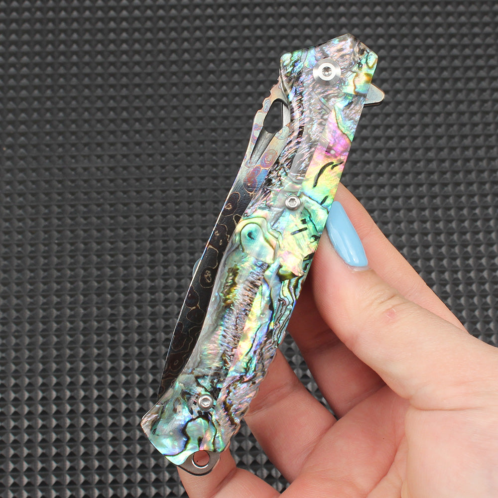 Close-up of a hand-held folded knife featuring a stunning abalone shell handle, displaying natural pearl-like iridescence and colorful textures, highly valuable for collection and decoration.