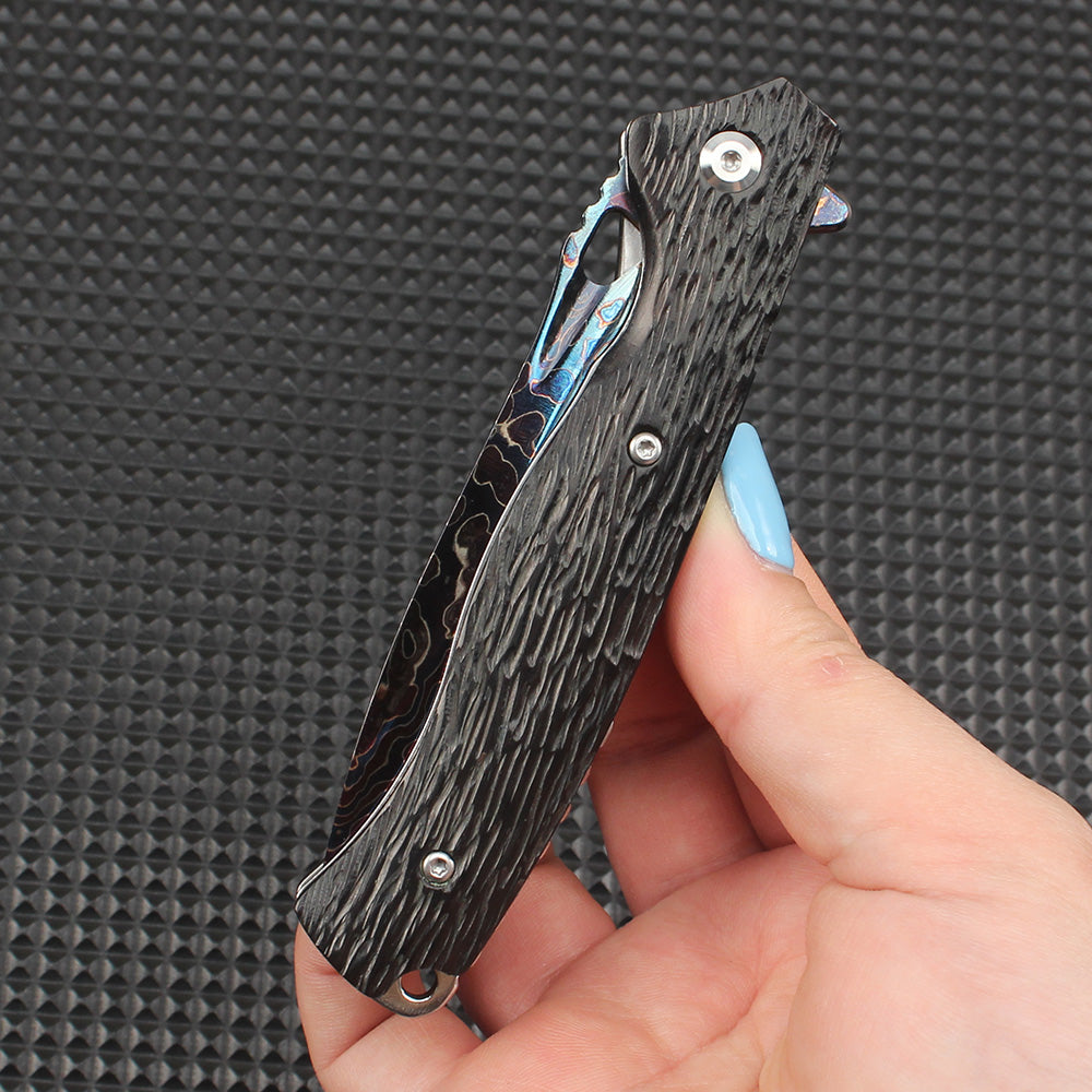 Hand-held display of this folding knife in the closed position, showing a rough bark-like textured dark handle for comfortable grip, equipped with a thumb stud and flipper tab.