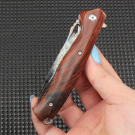 Close-up of a hand-held redwood handle folding knife in the closed position, showing natural wood grain and an oily sheen, highlighting the comfortable grip and design details.