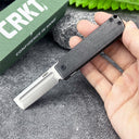 A hand holding an open CRKT folding knife with a black handle and silver tanto blade, displayed in front of a green CRKT box on a textured grey surface.