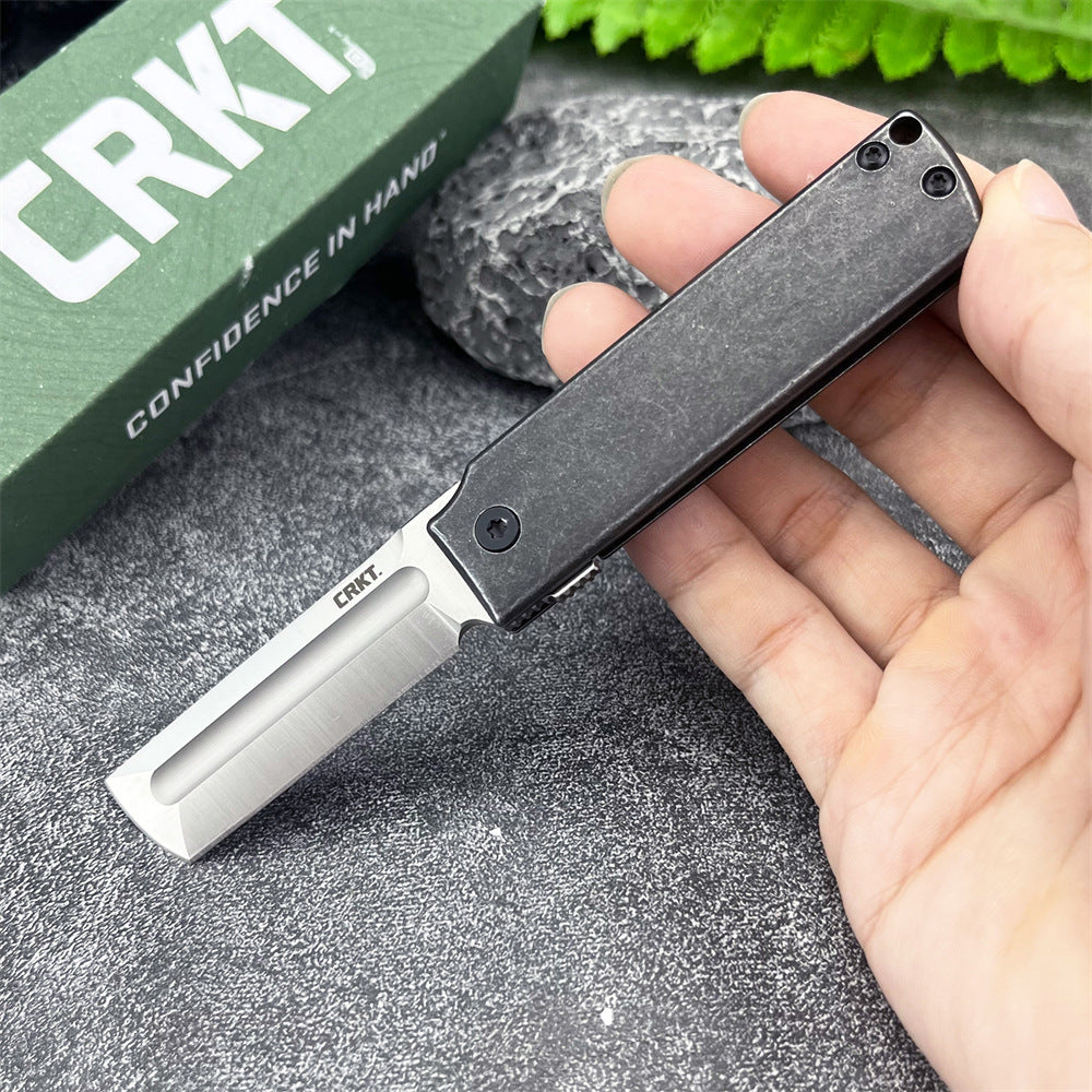 A hand holding an open CRKT folding knife with a black handle and silver tanto blade, displayed in front of a green CRKT box on a textured grey surface.