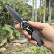 A hand holding an open folding knife with a black blade and gear pivot, displayed against an outdoor background with palm trees.