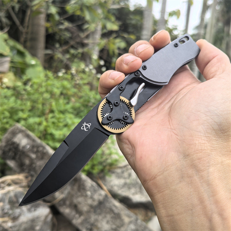 A hand holding an open folding knife with a black blade and gear pivot, displayed against a rocky outdoor background.