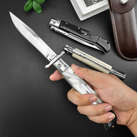 A hand holding an open Italian stiletto knife with a pearl white handle, resting on a dark leather surface. Two closed stilettos with black and bone handles and a brown leather sheath are visible in the background.