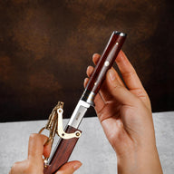 Hands demonstrating a folding knife with an M390 steel blade, a wood handle, and its matching wood sheath featuring a brass linkage and key ring, against a dark textured background.