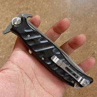 A hand holding a closed Noks folding knife, showing the black handle and silver pocket clip on a light wooden background.