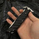 A hand holding a closed Noks folding knife, showing the black textured handle on a dark rocky background.