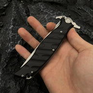 A hand holding a closed Noks folding knife, showing the black textured handle on a dark rocky background.