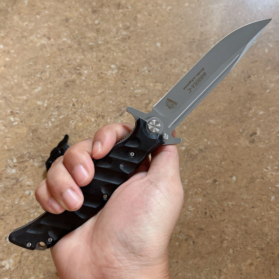 A hand holding an open Noks folding knife with a silver blade, displayed on a light wooden background.