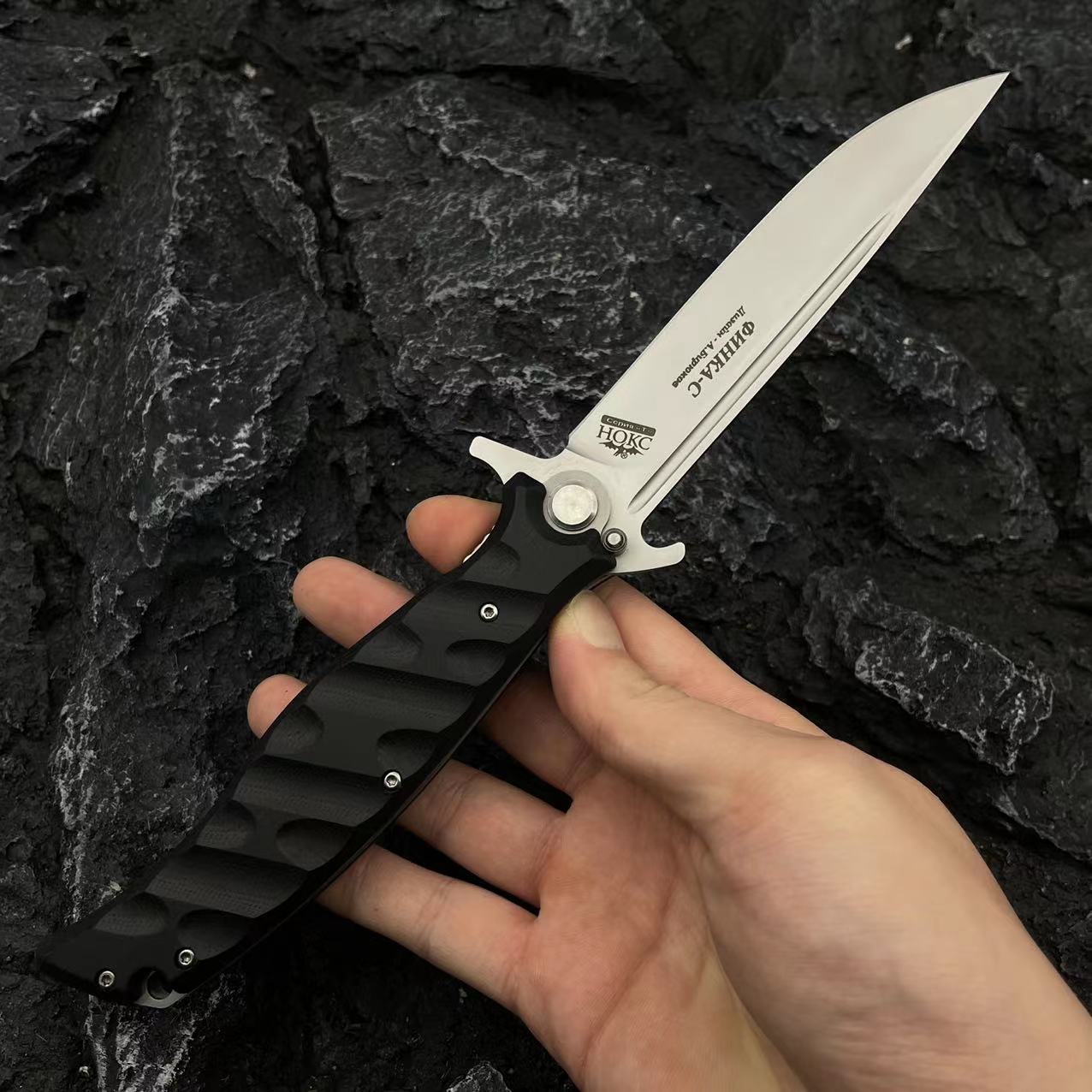 A hand holding an open Noks folding knife with a silver blade and black handle, displayed on a dark rocky background.
