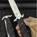 A hand wearing a tactical glove holding an open Noks folding knife, with a closed knife next to it on a black rope-textured surface.