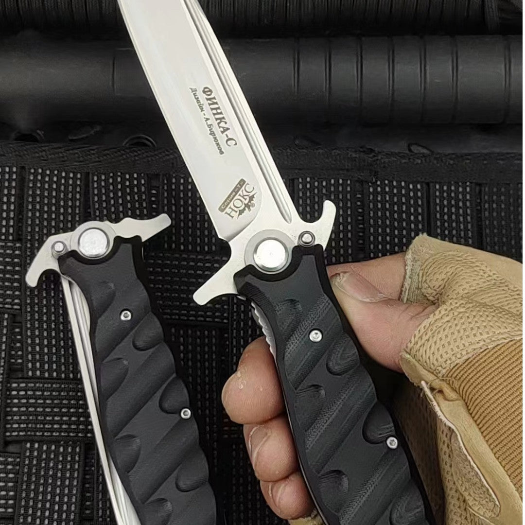 A hand wearing a tactical glove holding an open Noks folding knife, with a closed knife next to it on a black rope-textured surface.