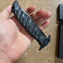 A bare hand holding a closed Noks folding knife, showing the side profile of the black textured handle, displayed on a light-colored textured surface.
