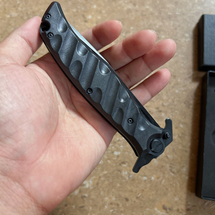 A bare hand holding a closed Noks folding knife, showing the side profile of the black textured handle, displayed on a light-colored textured surface.