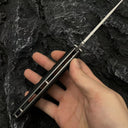 A bare hand holding an open Noks folding knife, showcasing the blade, side profile of the handle, and thickness, displayed on a dark rocky background.