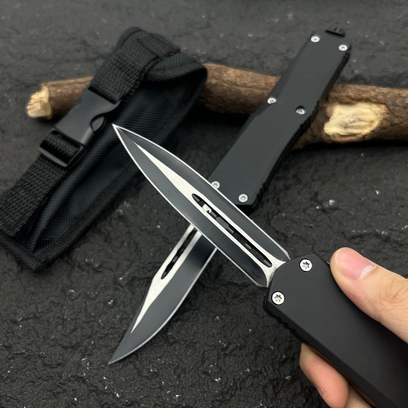 A hand holding a black OTF knife with a silver blade, with another black knife and a nylon sheath resting on a dark textured surface next to wood.