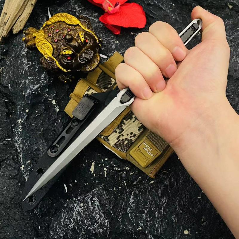 A hand gripping an open slim tactical folding knife, displayed with a black sheath, digital camo pouch, golden Buddha mask, and a red flower on a rough rock surface.