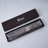 An unboxing shot of a JCHNC brand M390 folding knife wrapped in bubble wrap, resting inside its black packaging box on a white surface.