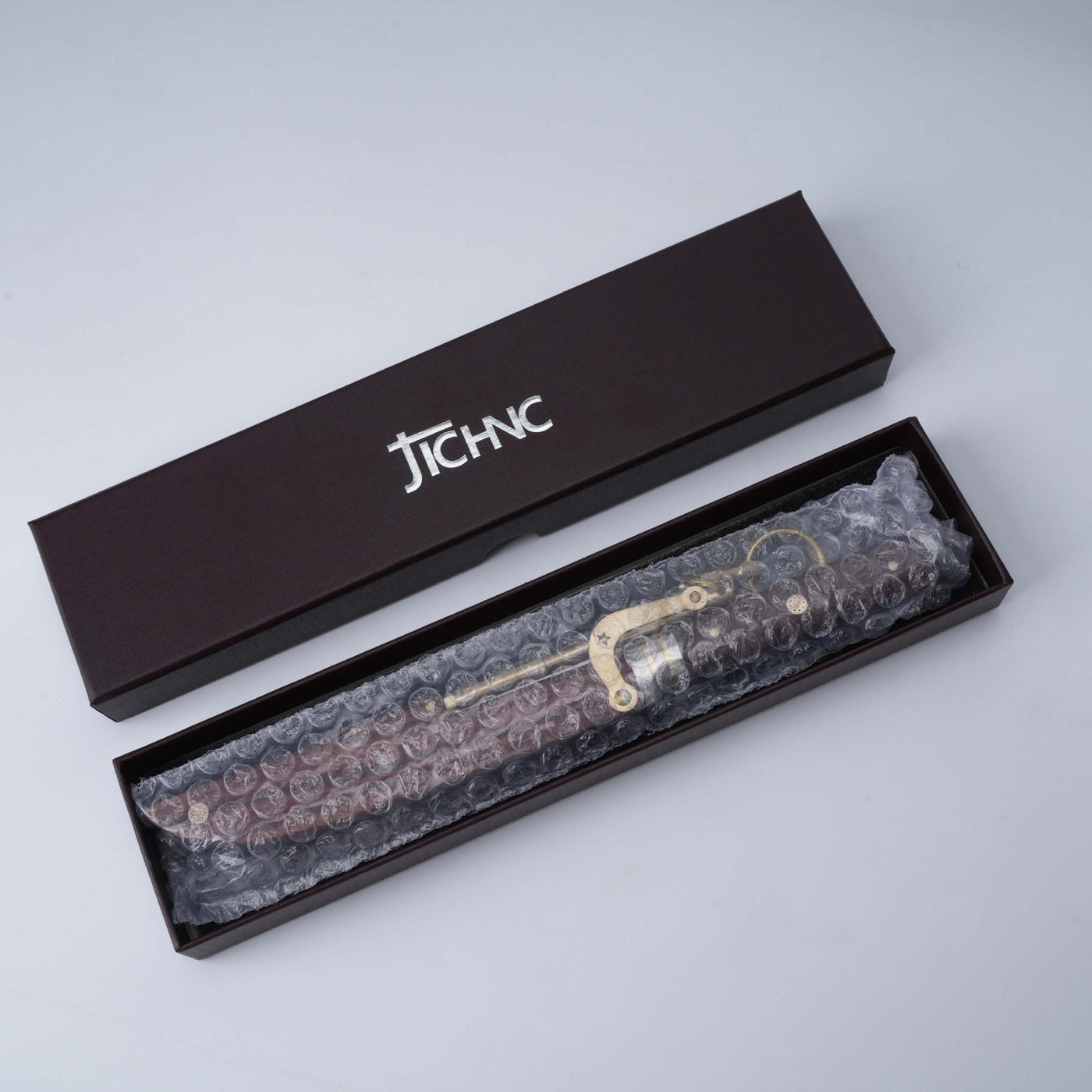 An unboxing shot of a JCHNC brand M390 folding knife wrapped in bubble wrap, resting inside its black packaging box on a white surface.