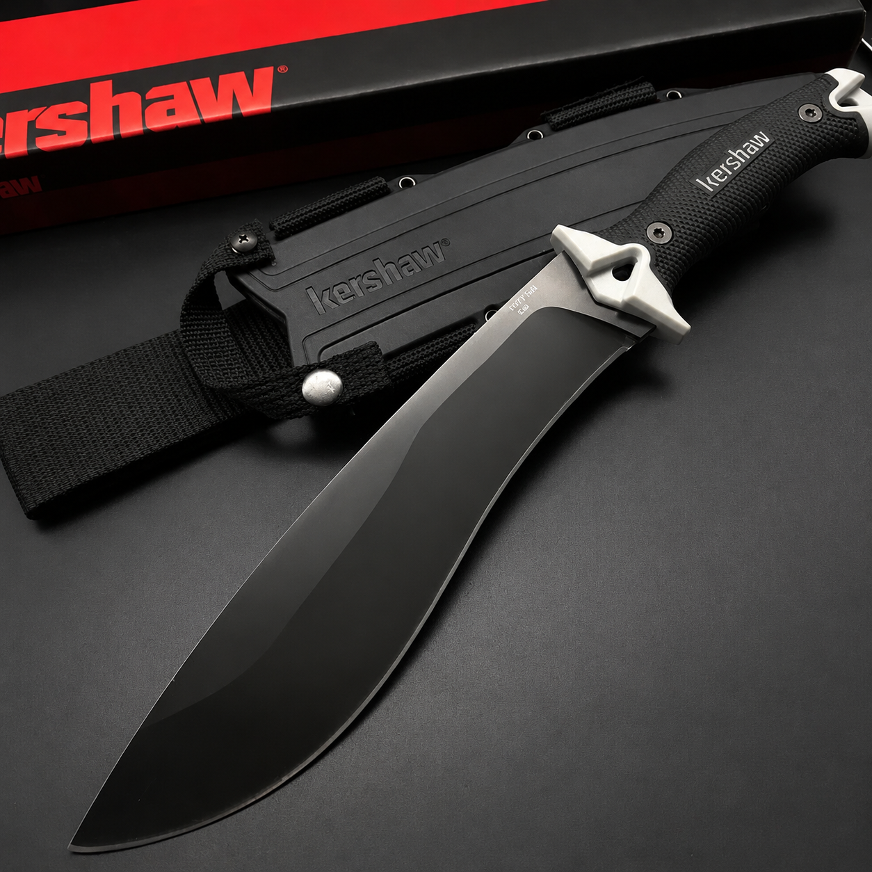 A Kershaw fixed blade knife with a full black blade and textured handle, featuring a silver guard, displayed next to its black molded sheath and red and black packaging box on a dark background.