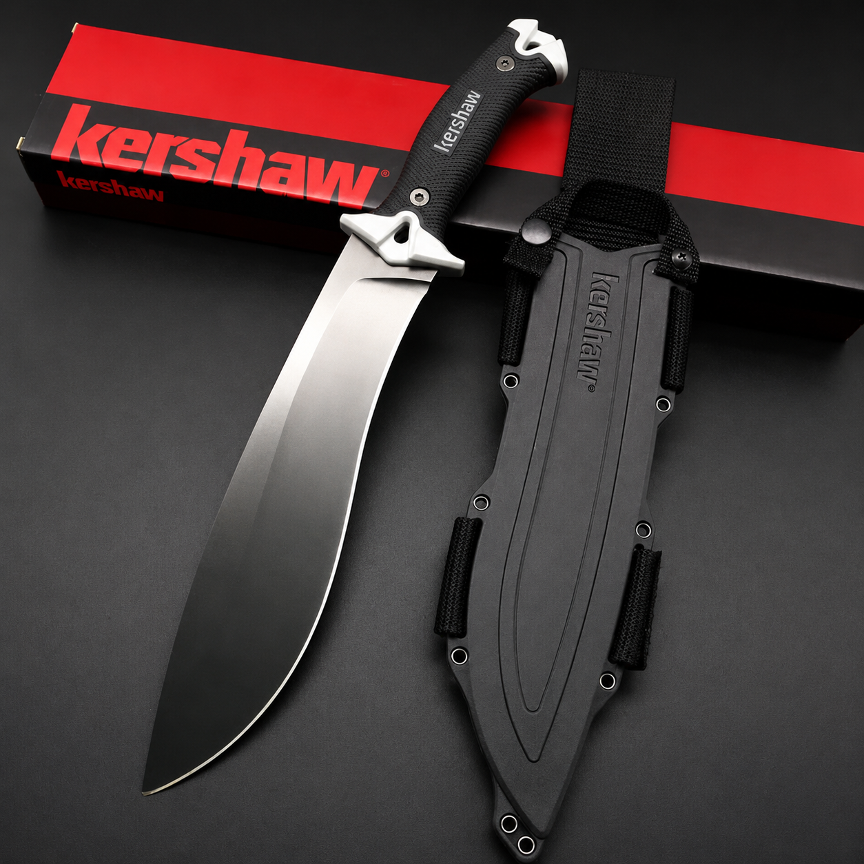 A Kershaw fixed blade knife with a black blade and textured handle, silver accents, displayed next to its black molded sheath and red packaging box on a dark background.