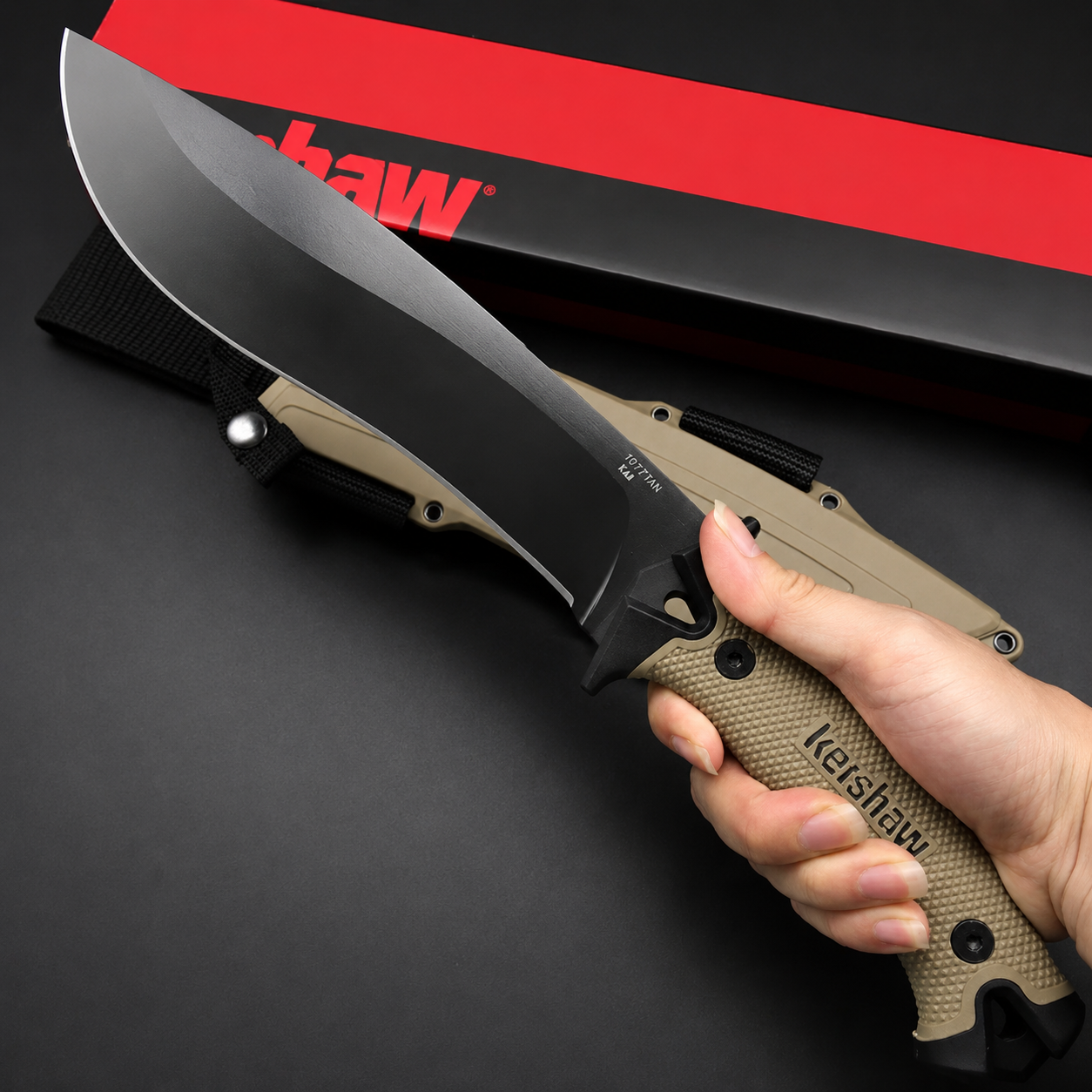A hand holding a Kershaw fixed blade knife with a tan textured handle and black blade, displayed above a tan sheath and red packaging box on a dark background.