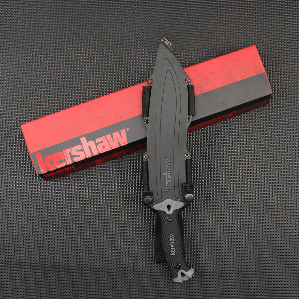 A sheathed Kershaw fixed blade knife with a black handle resting vertically on a red and black Kershaw packaging box, displayed on a dark gray textured surface.
