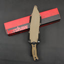 A top-down view of a sheathed Kershaw fixed blade knife with a tan handle and molded sheath, resting vertically across a red and black Kershaw packaging box on a dark textured surface.