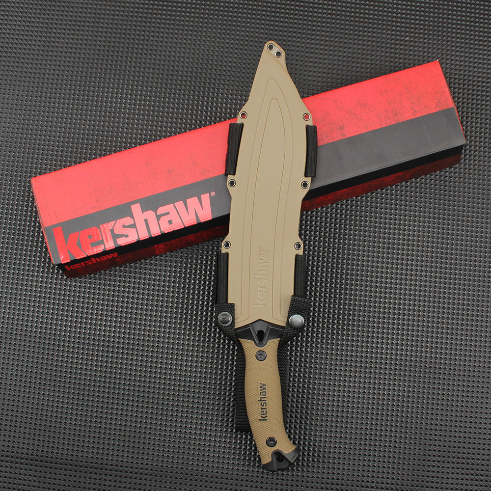 A top-down view of a sheathed Kershaw fixed blade knife with a tan handle and molded sheath, resting vertically across a red and black Kershaw packaging box on a dark textured surface.