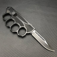 A view of the backside of a black knuckle duster folding knife, showing the textured inner holes of the knuckle guard and a black blade, on a carbon fiber background.