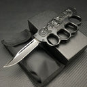 An open black knuckle duster folding knife displayed with its black nylon carrying sheath and black presentation box, resting on a carbon fiber textured background.