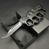 An open black knuckle duster folding knife displayed with its black nylon carrying sheath and black presentation box, resting on a carbon fiber textured background.