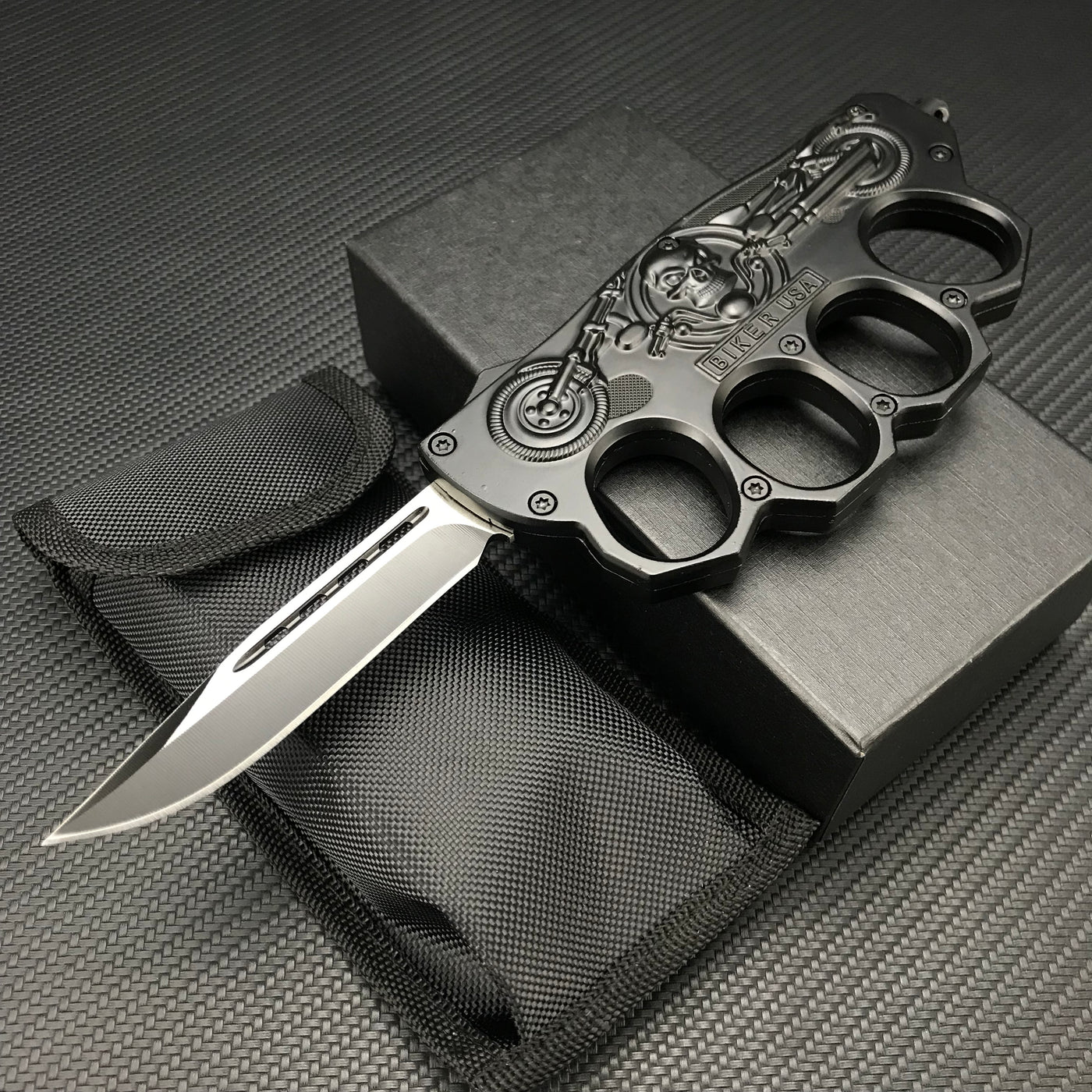 An open black knuckle duster folding knife displayed with its black nylon carrying sheath and black presentation box, resting on a carbon fiber textured background.