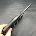 A hand holding an open black knuckle duster folding knife, showing the slender silver blade, the firing button, and the side profile of the handle, on a carbon fiber background.