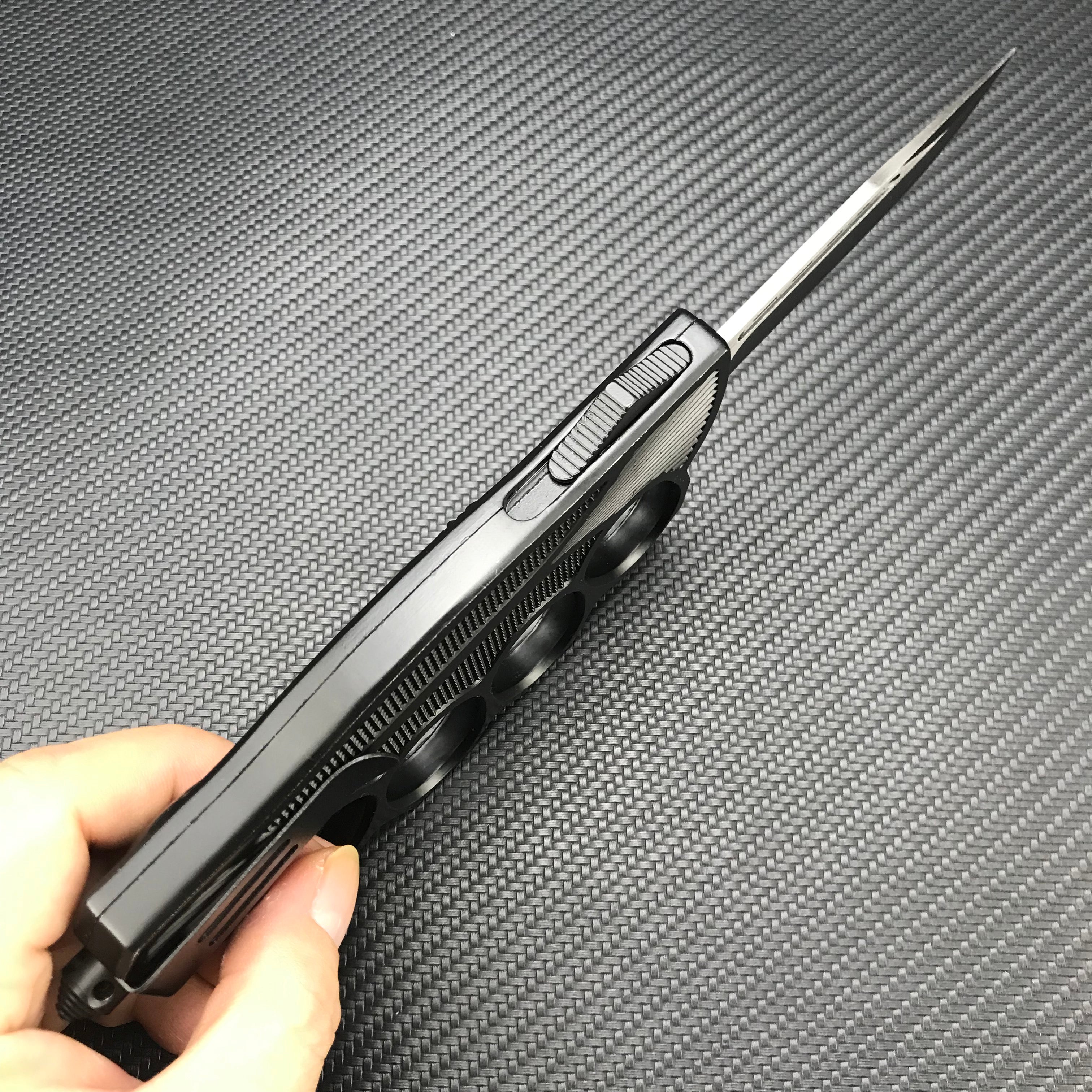 A hand holding an open black knuckle duster folding knife, showing the slender silver blade, the firing button, and the side profile of the handle, on a carbon fiber background.