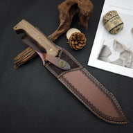 A handmade leather knife sheath with a wooden handle knife, displayed on a desk with pine cones, hemp rope, and decorative wood.