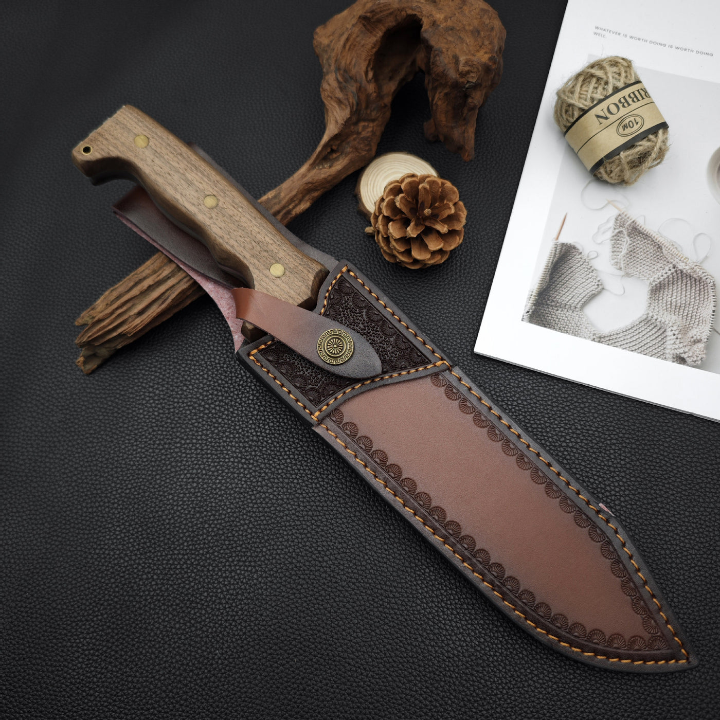 A handmade leather knife sheath with a wooden handle knife, displayed on a desk with pine cones, hemp rope, and decorative wood.