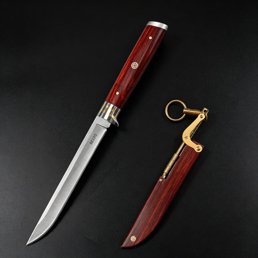An open M390 folding knife with a wood handle and metal guard, positioned next to its separate wooden sheath with a brass linkage and key ring, on a dark background.