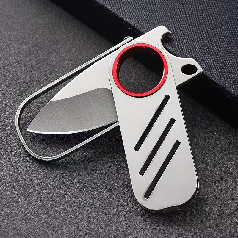 A close-up of a partially open EDC folding knife, showcasing its silver steel blade, red ring, and compact slide mechanism on a dark fabric background.