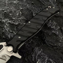 A close-up of the black textured handle and silver crossguard of a Noks folding knife on a rocky background.