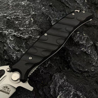 A close-up of the black textured handle and silver crossguard of a Noks folding knife on a rocky background.
