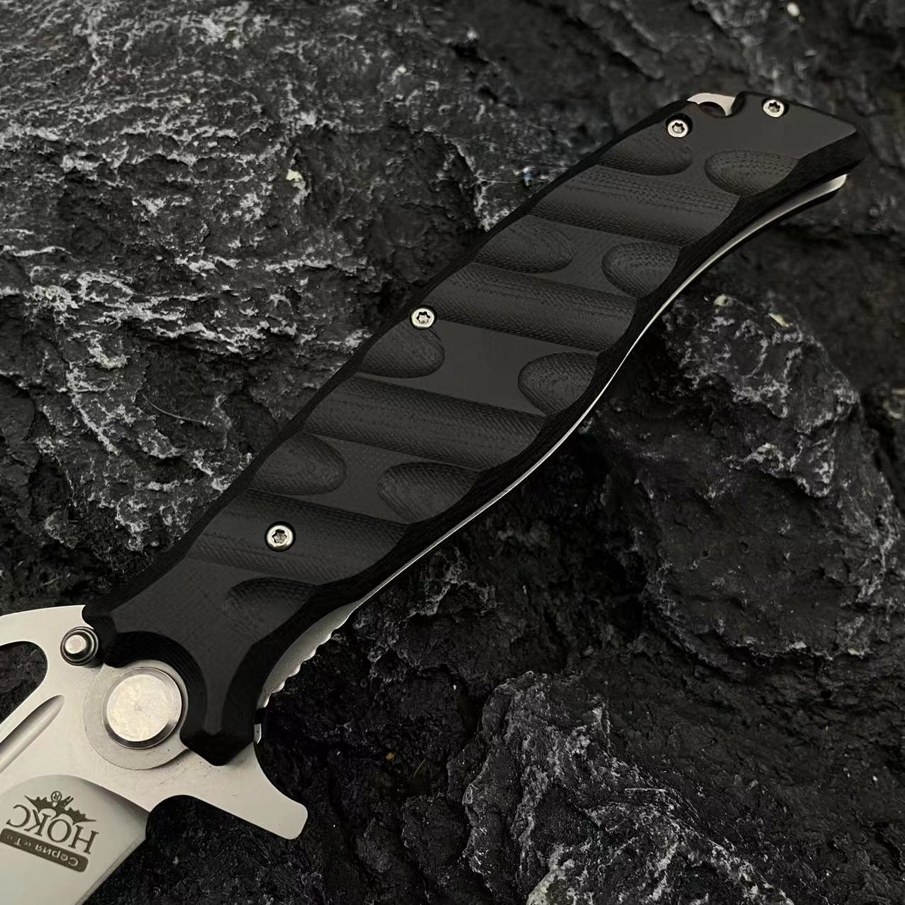 A close-up of the black textured handle and silver crossguard of a Noks folding knife on a rocky background.