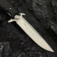 A close-up of the silver blade of a Noks folding knife showing the D2 steel stamp and fullers on a dark rocky surface.