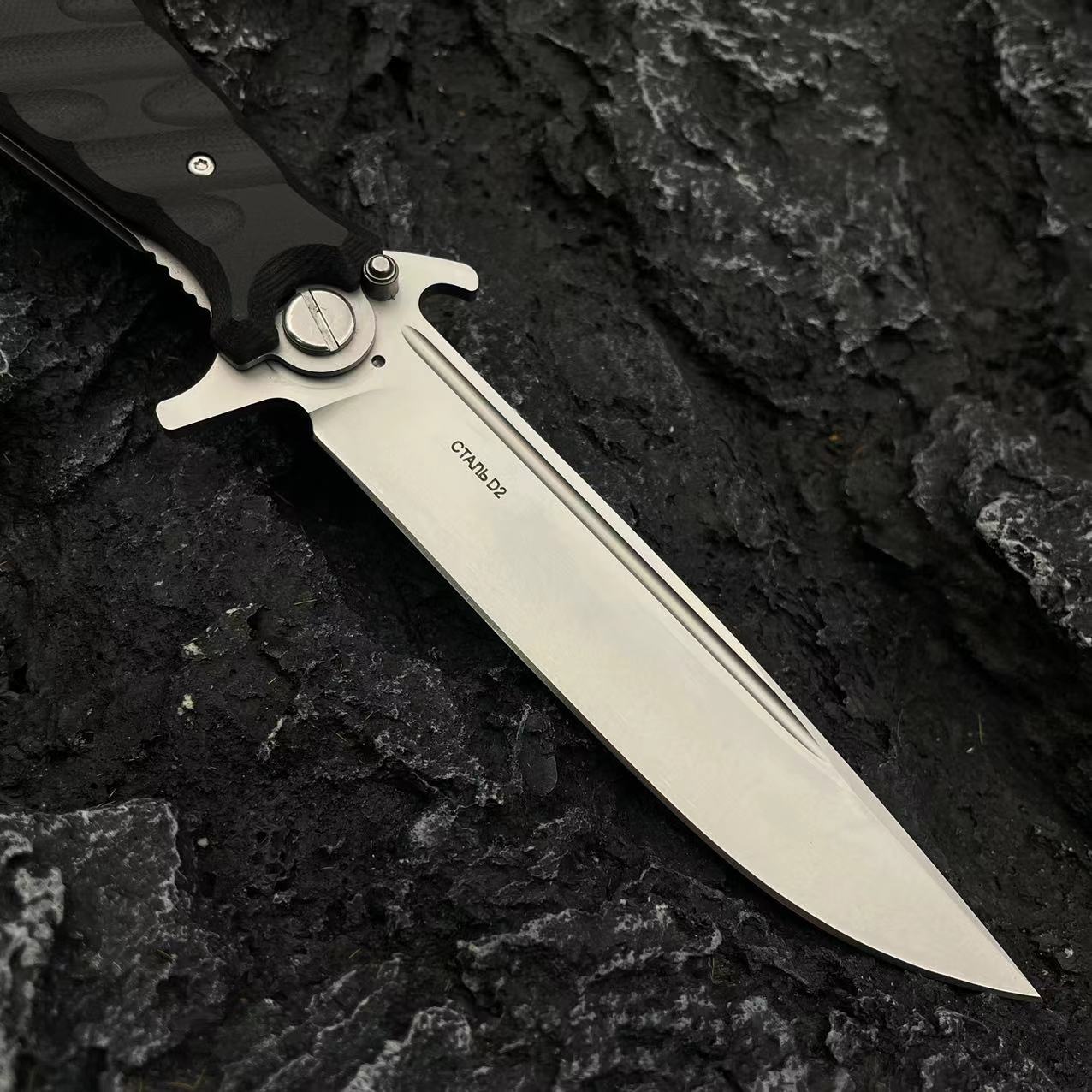 A close-up of the silver blade of a Noks folding knife showing the D2 steel stamp and fullers on a dark rocky surface.