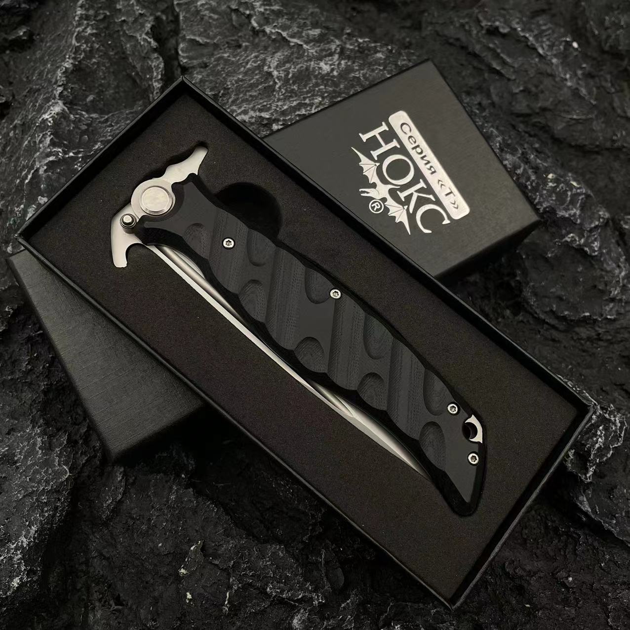 A Noks folding knife resting inside its black presentation box, displayed on a dark rocky background.