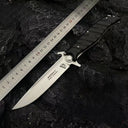 An open Noks folding knife with a silver blade, displayed on a rough rocky background next to a metal ruler for size reference.