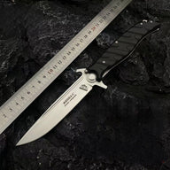 An open Noks folding knife with a silver blade, displayed on a rough rocky background next to a metal ruler for size reference.