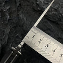 A close-up of the spine and thickness of a Noks folding knife blade, with a metal ruler measuring it on a dark rocky surface.