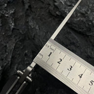 A close-up of the spine and thickness of a Noks folding knife blade, with a metal ruler measuring it on a dark rocky surface.