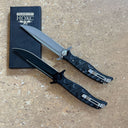 Two Noks folding knives with a black box on a light wooden surface. The top knife has a silver blade, while the bottom knife has a black blade.